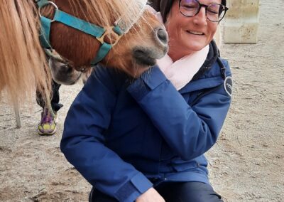 Melanie Preuß lacht herzlich, während ein kleines Pony sie sanft mit der Schnauze berührt – ein Moment voller Freude, Vertrauen und Harmonie zwischen Mensch und Pferd